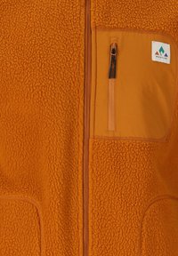Veste en polaire orange avec un tissu texturé, une poche poitrine zippée et un panneau lisse contrastant avec un ecusson logo.