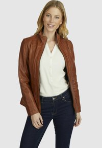 Giubbotto in pelle marrone con un design aderente, chiusura con zip, dettagli a costine e una texture morbida, indossato sopra una camicia leggera con bottoni e jeans scuri.