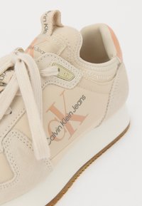 Beżowe sneakersy wykonane z zamszu i materiału tekstylnego, z logo "Calvin Klein Jeans" oraz subtelnym pomarańczowym akcentem na pięcie. Płaski design sznurowadeł.