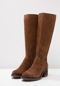 Botas de ante marrón hasta la rodilla con punta redonda y un tacón apilado. Presentan una costura vertical y una textura suave en el eje.