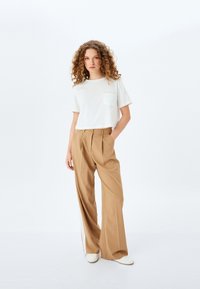 Camiseta blanca de manga corta con bolsillo, combinada con pantalones de pierna ancha de talle alto en color marrón claro. El modelo lleva zapatos blancos. Fondo minimalista.