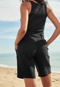 Black tank top paired with dark shorts. Shorts feature side pockets and a smooth texture, designed with a tailored fit. Background shows sand and sea.
