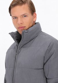 Grå pufferjakke med høj krave, der har en frontlynlås og quiltet design. Materialet ser let og glat ud.