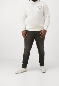 Uomo che indossa una felpa bianca con il logo "Hilfiger New York", jeans skinny grigio scuro e sneakers alte bianche, in piedi contro uno sfondo bianco.