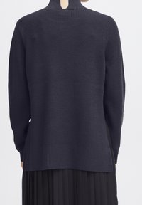 Pull en maille navy foncé avec un col montant, texture côtelée et fentes latérales, porté sur une jupe plissée noire.