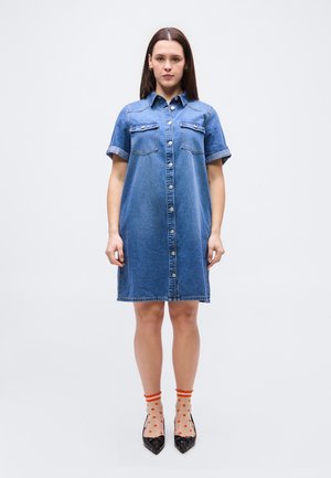 Egy nő áll előre nézve, kék farmerből készült, begombolt, felhajtott ujjú ruhát visel, narancssárga pöttyös áttetsző zoknival és fekete, hegyes orrú magassarkú cipővel.