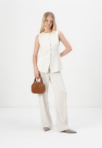 Gilet di lino chiaro con chiusura a bottoni, abbinato a pantaloni larghi. Una borsa marrone plissettata e delle scarpe beige con punta appuntita completano il look.