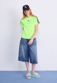 Νεόν πράσινη αθλητική μπλούζα κοντομάνικη με μαύρες ρίγες, σε συνδυασμό με ανοιχτό μπλε τζιν κολότ και μαλακές μίντ πράσινες παντόφλες. Μαύρο καπέλο.