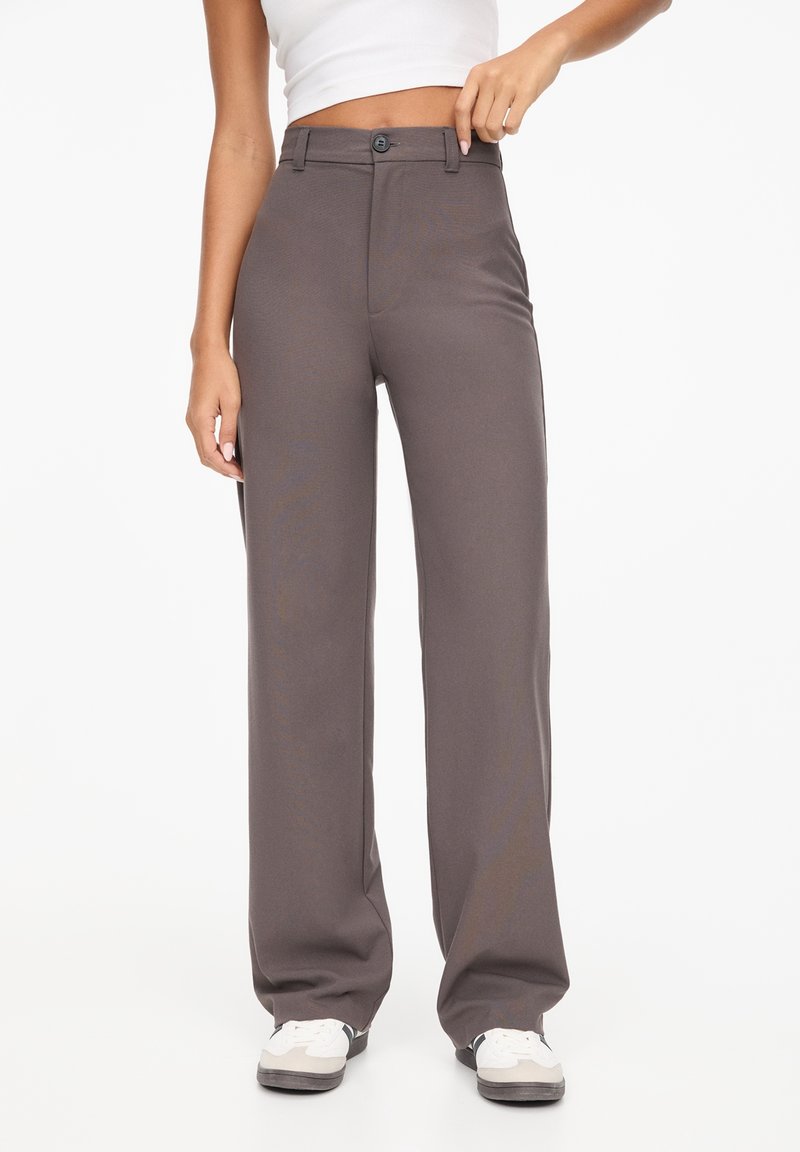 Personne portant un pantalon gris taille haute à jambes larges avec fermeture par boutons et des baskets blanches, debout devant un fond blanc uni.