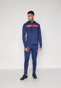 Conjunto de treino azul-marinho com capuz, apresentando painéis vermelhos e azuis na parte superior. Riscas brancas da Adidas ao longo dos lados das calças e detalhes com o logótipo.