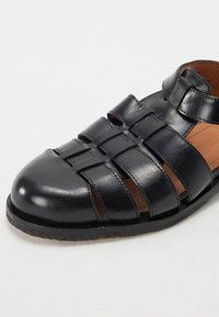 Black leather sandal with woven design, rounded toe, adjustable strap, and brown leather interior. Flat sole with slight texture.