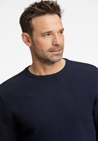 Laivastonsininen, teksturoitu neulepusero, jossa pyöreä pääntie. Kankaassa on hienovarainen neulottu kuvio. Mallilla on lyhyet hiukset ja kevyt parta.