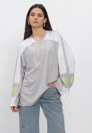 Vrouw draagt een oversized lichtgrijs mesh shirt met lange mouwen en gele letters, gecombineerd met losse blauwe spijkerbroeken, staand tegen een effen achtergrond.