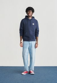 Ναυτικό φούτερ με κουκούλα και μπροστινή τσέπη, με λογότυπο, σε συνδυασμό με φαρδιά τζιν παντελόνια ανοιχτού μπλε χρώματος και κόκκινα αθλητικά παπούτσια. Μονόχρωμα, casual σχεδιασμός.