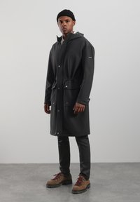 Długa czarna wodoodporna kurtka z kapturem, taliowana w pasie, z kieszeniami z przodu. Noszona na czarnych spodniach i brązowych, solidnych butach. Gładka faktura.