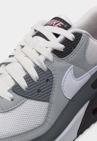 Tuva tuvs pelēkas un baltas krāsas Nike sporta kurpes ar baltām auklām un rozā "Nike" logotipu uz mēlītes.