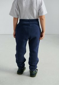 Tumši zilas vilnas sporta bikses ar elastīgu jostasvietu, šauru piegriezumu un potīšu aprocēm, valkātas kopā ar zaļām sandales. No aizmugures redzama pelēka T-krekla daļa.