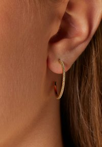 Gouden doorlopende oorbel met een gladde, glanzende afwerking en kleine, heldere rhinestones langs de buitenrand, hangend aan de oorlel.