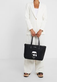 Svart axelväska med slät yta, med en tecknad figur i vitt, solglasögon och dragkedjestängning. Långa svarta handtag för bärande.