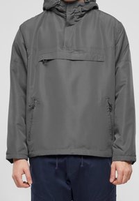 Man som bär en grå huva-jacka med framfickor och marinblå byxor, synliga från mitten av låret ner till händerna.