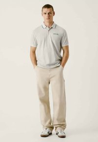 Ανοιχτό γκρι πόλο μπλουζάκι με αντίθετο γιακά, συνδυασμένο με μπεζ παντελόνι και λευκά αθλητικά παπούτσια. Ελάχιστο λογότυπο στη μπλούζα.