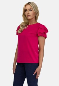 Fuksjowa bluzka z falbankowymi rękawami, krótka i dopasowana, gładka tekstura. W zestawie z wzorzystymi granatowymi spodniami. Prosty dekolt.