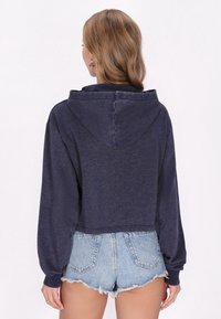 Sudadera recortada en azul marino, de tela suave, con mangas largas y amplias y una gran capucha; combinada con pantalones cortos de mezclilla azul claro.