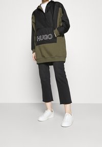 Huppari, jossa on musta yläosa ja vihreä alaosa, jossa on näkyvä "HUGO"-logolla varustettu tasku. Yhdistetty mustien 3/4-housujen ja valkoisten tennareiden kanssa.