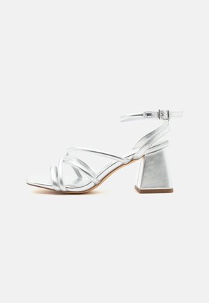 Sandale argentée à brides avec talon bloc et bride à boucle à la cheville, bout ouvert et semelle lisse sur fond blanc uni.