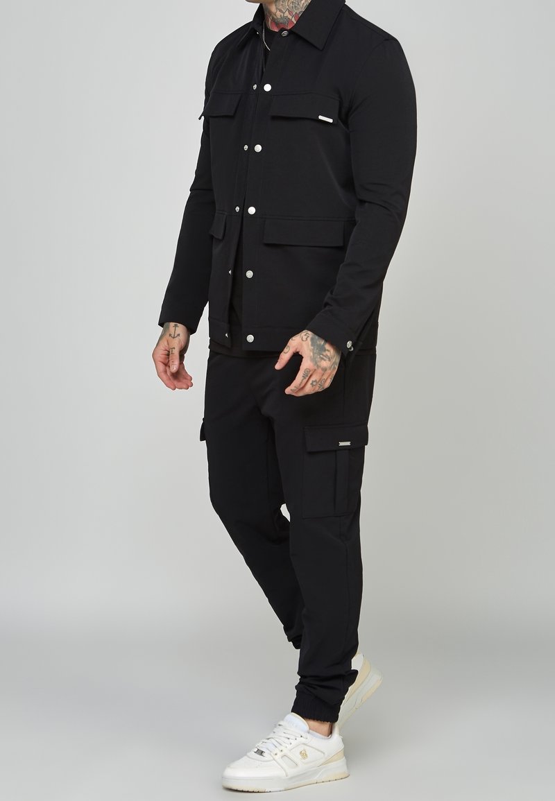 Homme portant une veste noire et un pantalon cargo avec des boutons argentés, des baskets blanches, et des tatouages visibles sur le cou et les mains.