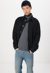 Chaqueta de mezclilla negra con botones plateados sobre una camiseta gris, combinada con jeans de color azul claro. Los accesorios incluyen un pañuelo negro con estampado.