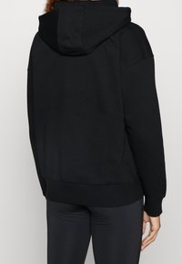 Persona che indossa una felpa con cappuccio nera semplice e pantaloni fitted neri, vista da dietro su uno sfondo chiaro e neutro.