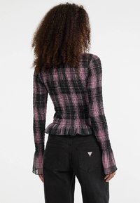 Frau mit lockigem Haar, die ein langärmliges Oberteil in Rosa und Schwarz mit strukturiertem Karomuster sowie schwarze Jeans trägt und mit dem Rücken vor einem weißen Hintergrund steht.