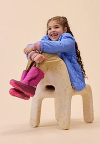Junges Mädchen mit langen, lockigen Haaren, das eine blaue Jacke, beige Hosen und rosa Stiefel trägt, sitzt und umarmt die Knie auf einem gesprenkelt kleinen Stuhl.