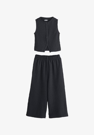 Zwart gestructureerd mouwloos topje met knopen, gecombineerd met bijpassende wijde broek met elastische taille, plat op een witte achtergrond gelegd.