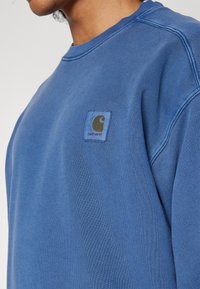 Sudadera de algodón azul con cuello redondo, puños acanalados y un pequeño parche de logo bordado marrón en el lado izquierdo del pecho. Tejido texturizado.