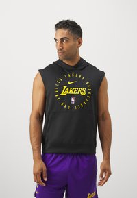 Melnā bezpiedurkņu kapuce ar dzelteno "Lakers" tekstu un logo. Iekļauj priekšējo kabatu un ar auklu regulējamu kapuci, kopā ar violetiem šortiem.