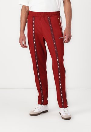 Homme portant un pantalon de survêtement Adidas rouge avec des points réfléchissants verticaux et des bandes blanches, associé à des baskets blanches et une chemise blanche.