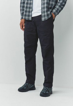 Man wearing black cargo pants, black and gray athletic shoes, a white shirt, and a gray checkered long-sleeve shirt, standing against a plain background.