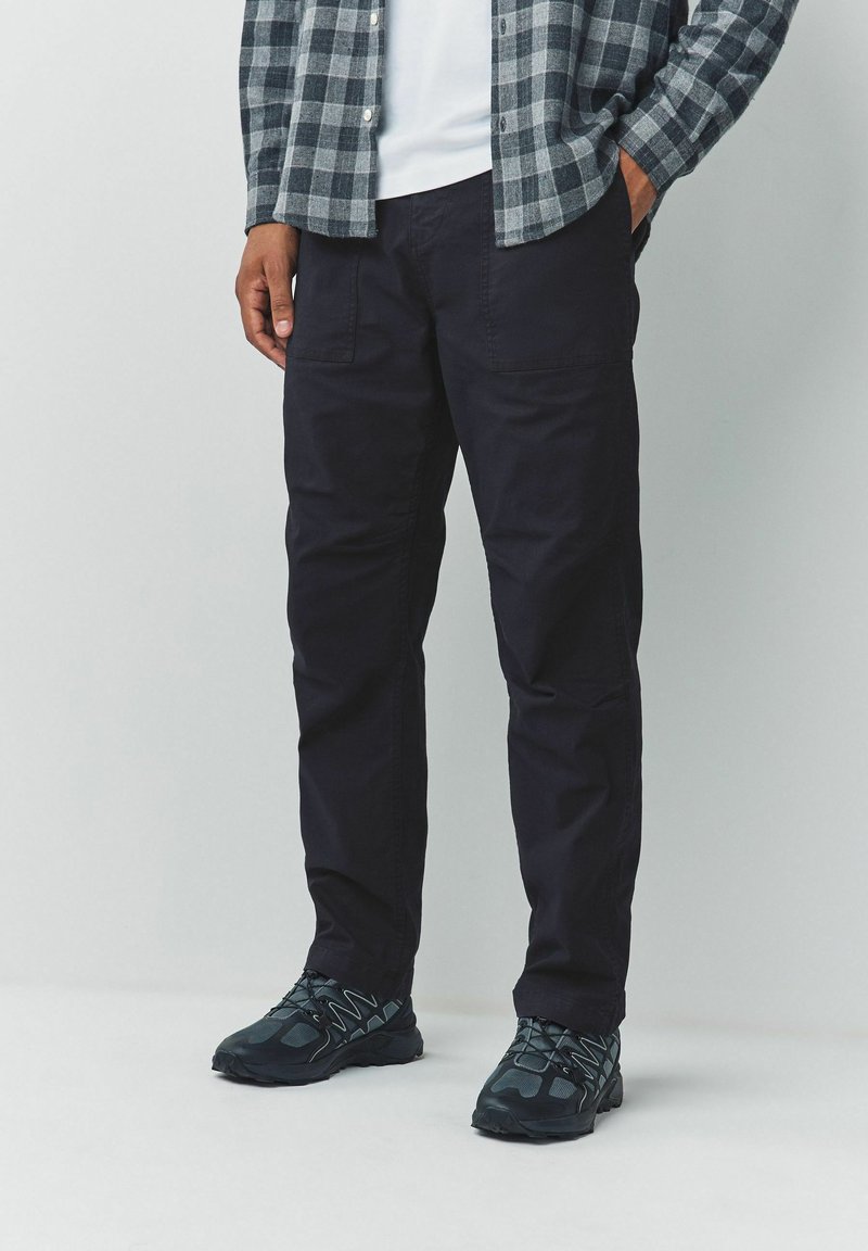 Man wearing black cargo pants, black and gray athletic shoes, a white shirt, and a gray checkered long-sleeve shirt, standing against a plain background.
