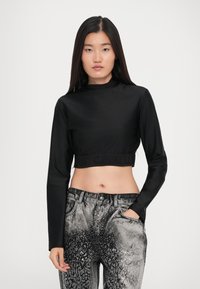 Young woman wearing a black long-sleeve crop top and gray high-waisted jeans with a white dotted pattern, standing against a white background.