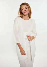 Cardigan aperto in morbido maglione bianco, con maniche ampie e una vestibilità morbida. Indossato su una maglietta chiara e pantaloni bianchi cintrati. Texture minimale.