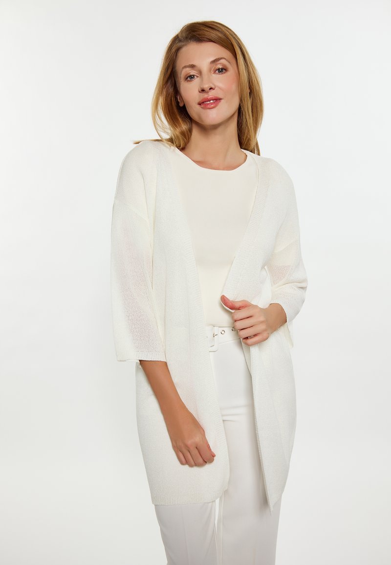 Cardigan aperto in morbido maglione bianco, con maniche ampie e una vestibilità morbida. Indossato su una maglietta chiara e pantaloni bianchi cintrati. Texture minimale.