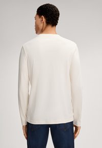 Langarmshirt aus cremefarbener Baumwolle mit Rundhalsausschnitt, glatter Textur und entspannter Passform, kombiniert mit dunkelblauen Jeans.