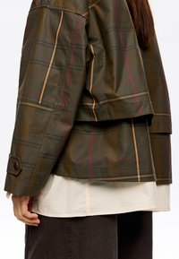 Kraciasta kurtka w ciemnozielonym kolorze z brązowymi, żółtymi i różowymi paskami. Charakteryzuje się prostokątnym kształtem, zapięciem na guziki przy mankietach i warstwowym dołem na beżowej koszuli.