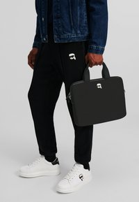 Geantă rectangulară neagră cu două mânere, fabricată din material sintetic, având un detaliu cu logo. Asortată cu pantaloni negri și teniși albi.