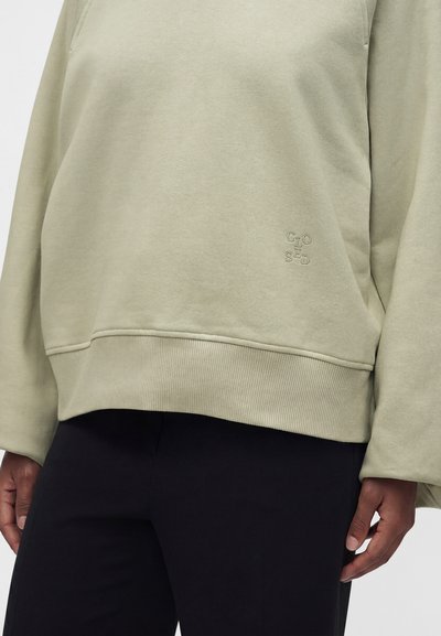 Personne portant un sweatshirt beige clair avec "CLOSED" brodé près de l’ourlet et un pantalon noir, vue des épaules aux cuisses.