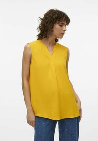 Top giallo senza maniche con scollatura a V, tessuto strutturato e vestibilità comoda, abbinato a jeans blu con piccoli ornamenti in argento.