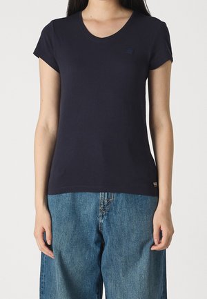 Femme portant un t-shirt uni bleu marine foncé à col en V et manches courtes ainsi qu'un jean bleu ample, debout sur un fond clair.
