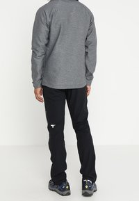 Haut gris à manches longues avec un col rond, associé à un pantalon noir avec une poche zippée sur le côté et un logo réfléchissant. Les chaussures sont sombres avec des accents vifs.